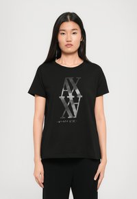 Pikkade tumedate juustega naine kannab musta Armani Exchange särk, millel on läikivad kattuvad "AX" tähed, ja musti pükse, seistes valge tausta ees.