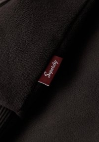 Tela de punto negra con una textura suave, con una etiqueta roja que tiene el texto blanco "Superdry" de un lado.