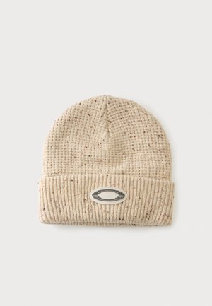 Bonnet tricoté beige avec fil moucheté et revers plié avec un écusson ovale brodé noir et blanc.