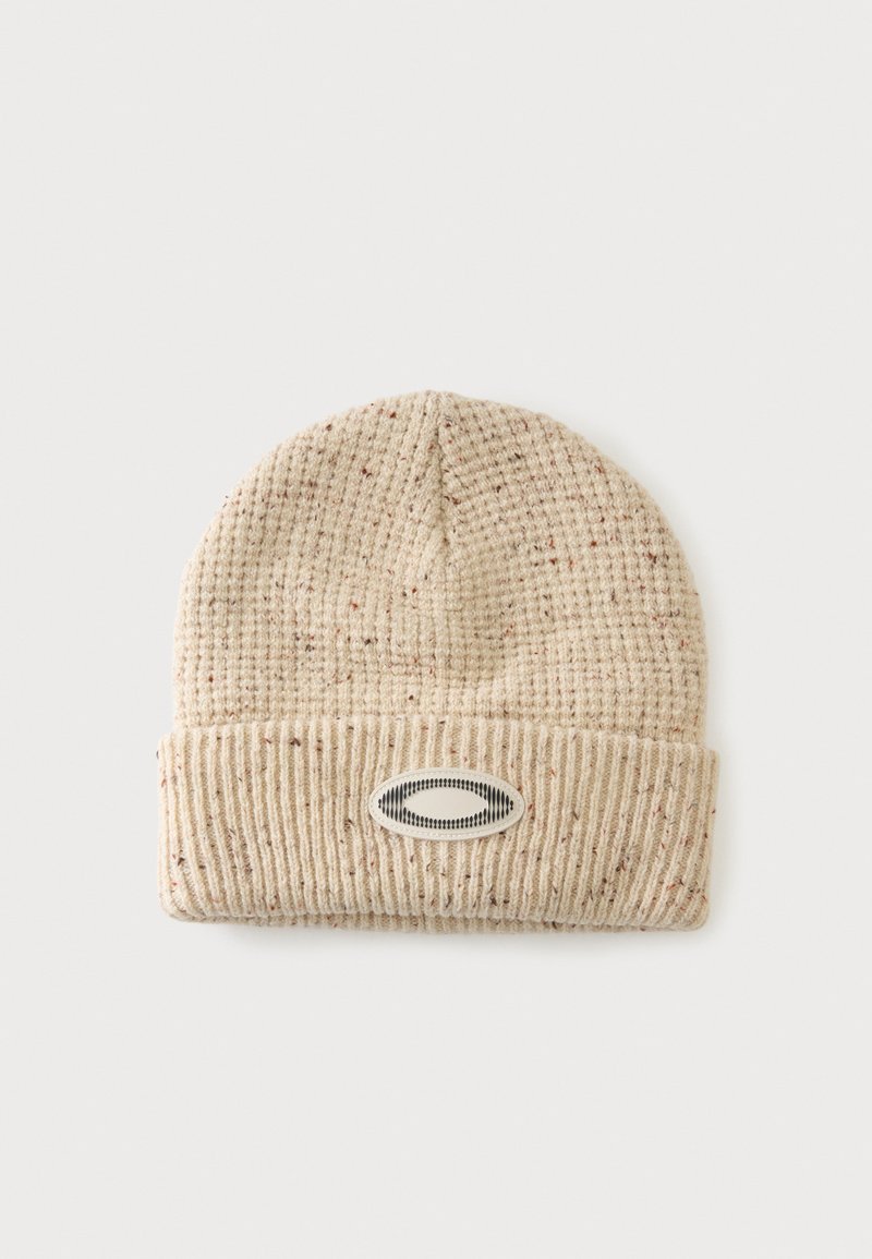 Bonnet tricoté beige avec fil moucheté et revers plié avec un écusson ovale brodé noir et blanc.