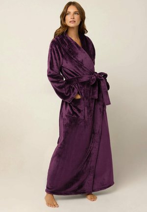 Femme portant un long peignoir en peluche violet foncé avec une ceinture à la taille et des poches, debout pieds nus devant un fond clair uni.