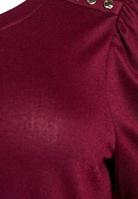 Burgunderfarbener Strickoberteil mit gerippter Textur, gerafften Ärmeldetails und drei metallischen Nieten an der Schulter als Akzent.
