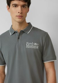 Grijze polo shirt van een gestructureerde stof, witte accenten op de kraag, en het geborduurde 'Ford Mustang' logo op de linkerkant van de borst.