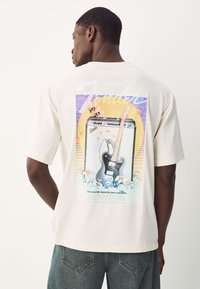 Cream-colored t-shirt featuring a colorful graphic of an amplifier and guitar with floral accents and playful penguin illustrations on the back.