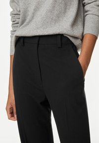 Zwarte op maat gemaakte broek van gladde stof, met een platte voorkant, zijzakken en riemlussen. Gecombineerd met een grijze gebreide trui.