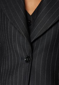 Czarna marynarka w prążki z klapami z wycięciem, pojedynczym zapięciem na guzik oraz teksturowaną tkaniną z kontrastującymi białymi paskami.