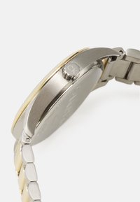 Reloj de pulsera de acero inoxidable con bisel acentuado en dorado, que presenta una caja redonda, una correa de eslabones metálicos y una corona etiquetada.