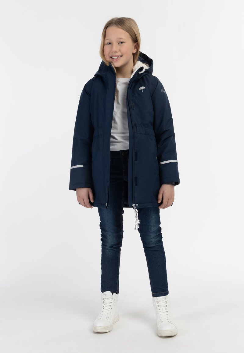 Giacca outdoor navy con cappuccio foderato morbido, chiusura con zip e vita elastica. Accenti bianchi e riflettori sulle maniche. Abbinata a jeans blu e sneaker bianche.