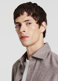 Camicia marrone con bottoni, dalla texture morbida, con colletto e patta con bottoni nascosta; indossata sopra una t-shirt bianca.