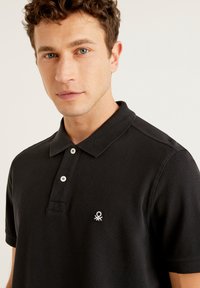 Černé polo tričko z texturované látky, s dvouknoflíkovým zapínáním a malým vyšitým logem na levé straně hrudi.