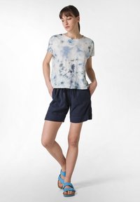 Světle modré tie-dye bavlněné tričko v kombinaci s tmavě modrými šortkami; obuv tvoří modré nastavitelné sandály. Čisté linie a uvolněný střih.