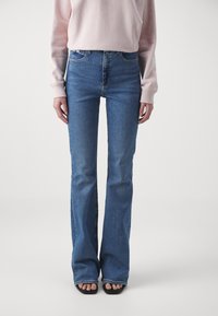 Vaqueros acampanados de mezclilla en azul, con una cintura alta, diseño de pierna recta y líneas limpias. Combinados con un sudadera rosa clara y sandalias negras.