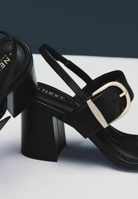 Schwarze Sandaletten mit Blockabsatz, Riemen aus Leder und einem glänzenden goldfarbenen Schnallenakzent. Glatte Textur und minimalistisches Design.