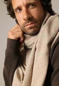 Écharpe beige tricotée drapée sur un pull marron foncé, présentant une texture douce et un motif subtil, portée de manière décontractée autour du cou.