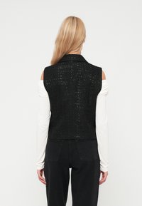 Crna skraćena majica s teksturiranim tkanjem, s ovratnikom i bez rukava, uparen s dugim rukavima bijele majice i crnim hlačama.