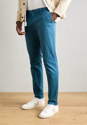 Mand iført slanke teal bukser, beige jakke, hvide sneakers med beige "V"-logo, stående på trægulv mod neutral baggrund.