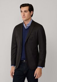 Blazer grigio scuro con rever a punta, chiusura a un bottone e due tasche frontali. Indossato sopra un maglione blu a scollo a v e una camicia azzurra chiara.