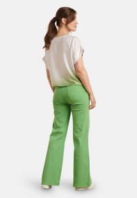 Green flared jeans with rear pockets, paired with a light, slightly sheer, short-sleeve white top. The fit is casual.