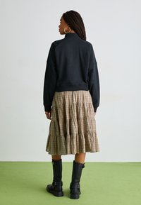Sudadera negra sin cuello combinada con una falda de cuadros en tonos beige y marrón con volantes. Botas negras altas hasta la rodilla con hebillas. Fondo verde.