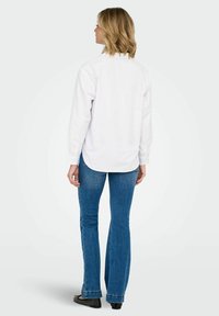 White long-sleeve shirt with a curved hem, paired with blue flared jeans and black shoes. Smooth fabric with practical design details.