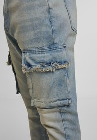 Jeans en denim bleu clair avec une poche cargo à bord effiloché sur la cuisse, mettant en valeur des coutures détaillées et la texture du tissu.