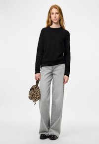 Jauna moteris su ilgais raudonais plaukais vilki juodą megztinį, šviesiai pilkas plačias kelnes, juodus batus ir laiko leopardo raštuotą rankinę.