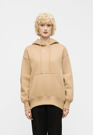 Personne aux cheveux blonds bouclés et courts, portant un sweat à capuche Nike beige et un pantalon noir, debout devant un fond blanc uni.