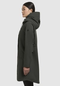 Lång mörkgrön regnrock med huva, med dragkedjestängning, sidofickor och en slät yta. Utsmyckad med en logotyp på ärmen.