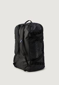 Sort Patagonia gymtas med skulderremme og håndtag, stående oprejst på en ensfarvet lys baggrund.