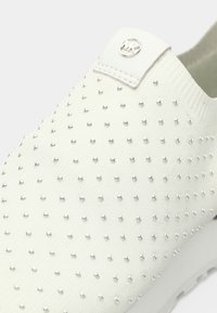 Chaussure blanche à enfiler avec un tissu texturé, ornée de petits clous argentés. Comprend une étiquette logo et une semelle extérieure flexible.