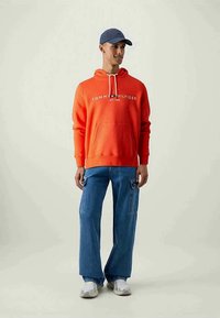 Sudadera naranja con bolsillo frontal y logo blanco. Pantalones de denim azul de corte holgado, con bolsillos de carga. El modelo lleva una gorra oscura y zapatillas blancas.
