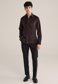 Camicia marrone con bottoni e un motivo testurizzato, abbinata a pantaloni neri su misura e mocassini neri, indossata da un modello in posizione eretta.