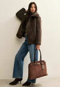 Kobieta w brązowej kurtce z owczej skóry, niebieskich dżinsach i czarnych szpilkach, trzymająca dużą brązową torebkę z zamszu ozdobioną zawieszkami.