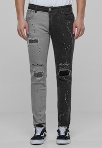 Falmede grå og svarte jeans med slitte detaljer og malingsprut aksenter. Har rifter og brodert merkevare langs sidene.