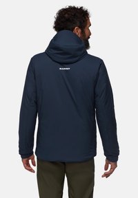 Marinblå isolerad jacka med huva, texturerad tyg och en vit logotyp på ryggen. Slät yta med elastiska manschetter och sid sömmar.