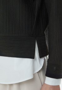 Black ribbed top with textured fabric, featuring a straight hem and subtle brand detailing on the sleeve. White shirt visible underneath.