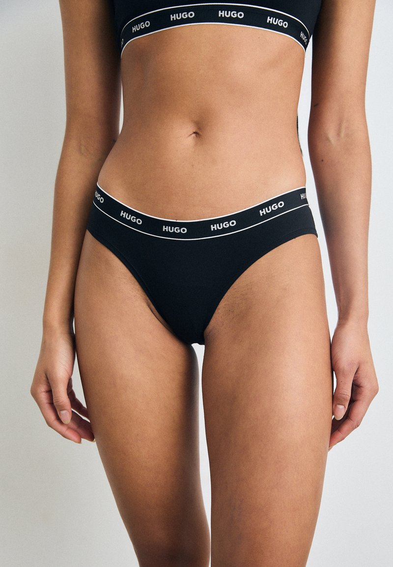 Costum de baie din bumbac negru, cu un design cu tăietură înaltă. Prezintă un brâu alb cu logo și o textură netedă. Stil minimalist, fără modele.