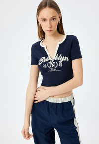Camiseta recortada azul marino con textura acanalada, escote en V abotonado y estampado gráfico blanco que dice "Brooklyn 21 18". Combinada con pantalones azul marino.