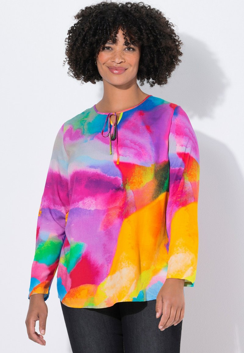 Vrouw met krullend haar, gekleed in een langemouwen blouse met een meerkleurig aquarelpatroon en donkere broek, die glimlacht tegen een witte achtergrond.