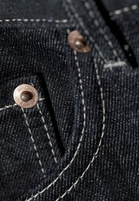Gros plan d’un tissu en denim noir avec des coutures blanches et des rivets en cuivre sur le détail d’une poche de jean.