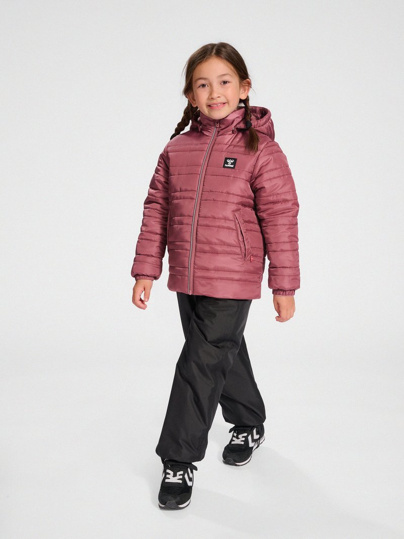 Kind met gevlochten haar draagt een mauve gewatteerde jas met capuchon, zwarte broek en zwarte sneakers, lopend in een lichte studio met een witte achtergrond.