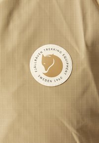 Beige fabric with a grid pattern. Features a circular logo patch in white and gold with a fox design, labeled "Fjällräven Trekking Equipment."