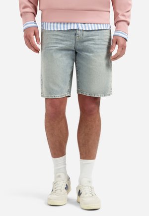 Man draagt lichtblauwe denim shorts met een roze sweatshirt, blauw gestreept shirt, witte sokken en witte sneakers.