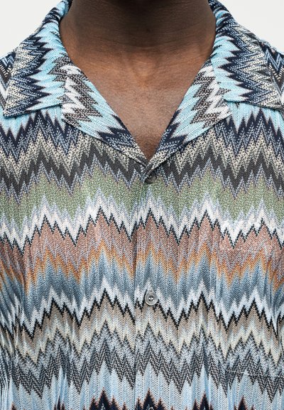 Κοντινό πλάνο ενός ατόμου που φοράει πουκάμισο με κουμπιά και πολύχρωμο ζιγκ ζαγκ μοτίβο σε αποχρώσεις του μπλε, πορτοκαλί, πράσινου, μαύρου και λευκού.