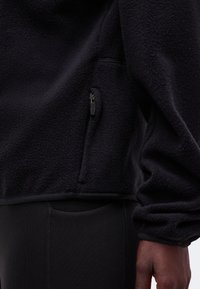 Veste polaire noire avec poche latérale zippée portée sur un pantalon noir ajusté, montrant le bas du torse et la main droite d'une personne.