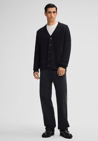 Czarny dzianinowy kardigan z dekoltem w serek, z teksturowanym wzorem i zapięciem na guziki, noszony na białej koszuli i zestawiony z czarnymi spodniami.