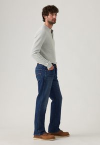 Camisa henley de manga larga gris, jeans de mezclilla azules y zapatos marrones. El modelo está de lado, con las manos en los bolsillos, mostrando el ajuste y el color de la ropa.