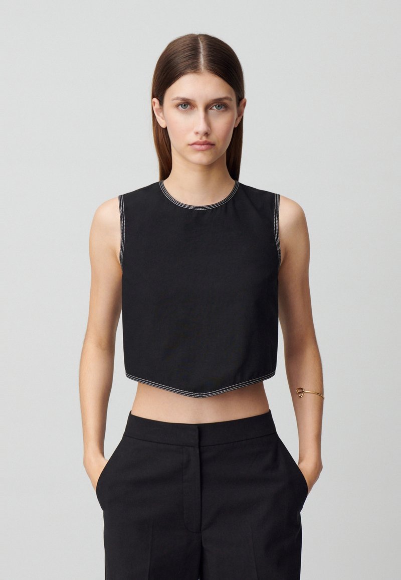 Top corto negro sin mangas con escote redondo y costuras blancas a lo largo de los bordes, combinado con pantalones negros de cintura alta.