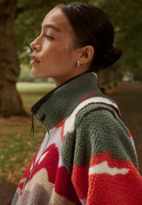 Pullover in fleece con un motivo astratto verde, rosso e bianco. Collo alto, zip frontale a metà e una finitura morbida e testurizzata.