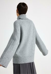 Person med kort, mørkt hår iført sølvøreringe og en løs, lys grå strik turtleneck sweater med brede ærmer og mørk nederdel.